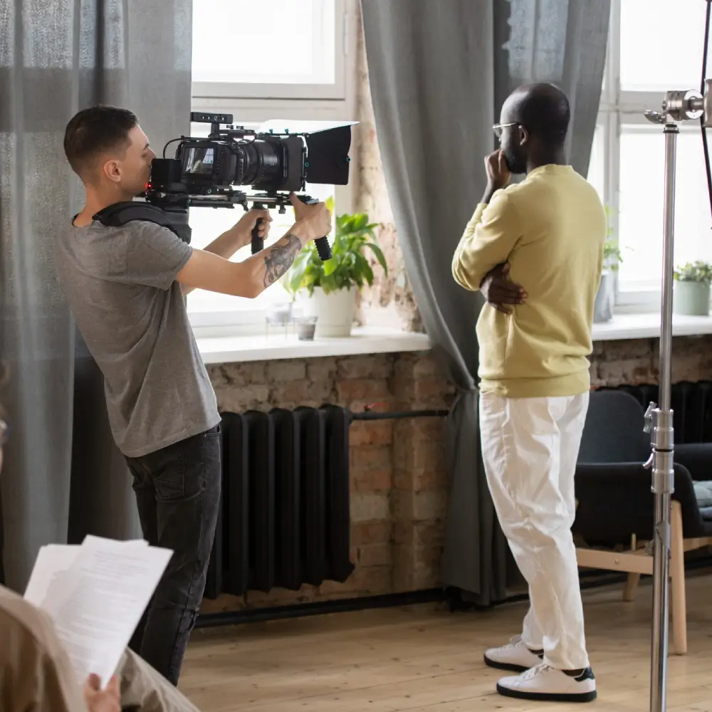 A cameraman filming a man standing by a window indoors.